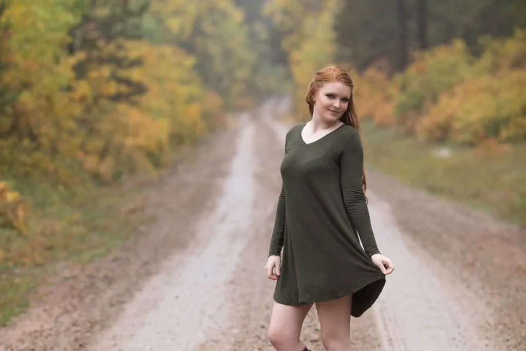 Młoda kobieta pozuje pewnie na polnej drodze otoczonej jesiennymi liśćmi.