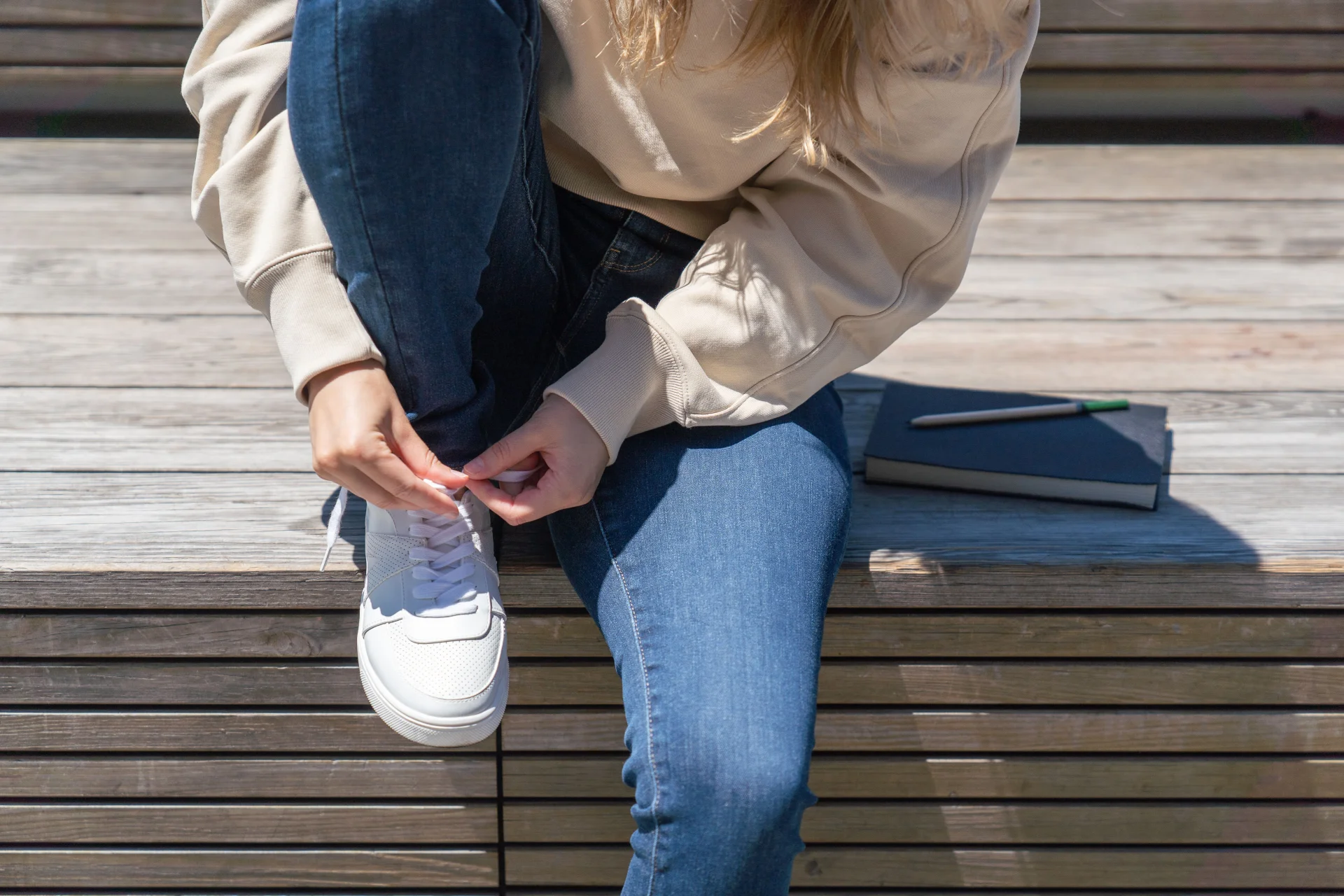 Kobieta w dżinsach zawiązuje białe sneakersy, siedząc na drewnianych schodach obok notatnika.